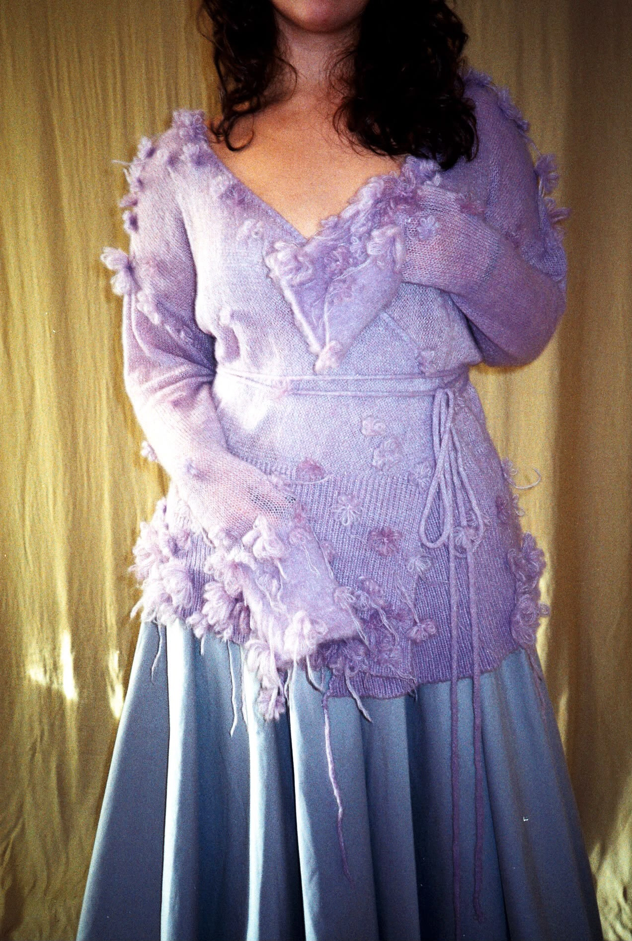 Model wearing a lilac handcrafted knitted cardigan with textured floral details over a light blue skirt, highlighting innovative knitwear design and artisanal craftsmanship. 